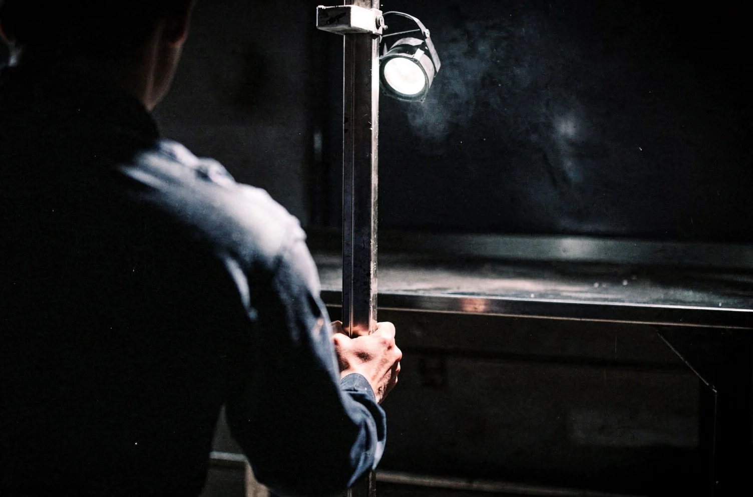 A person seen from behind holding the upright of a heavy steel door under a single work lamp, the bench behind them empty