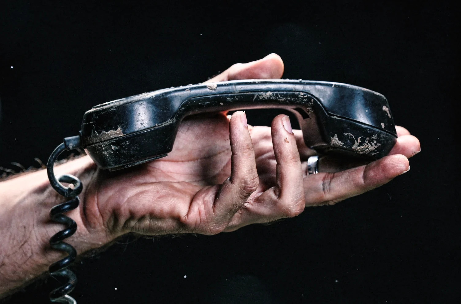 A hand holding a battered black corded telephone handset in an open palm, dust settled in the seams