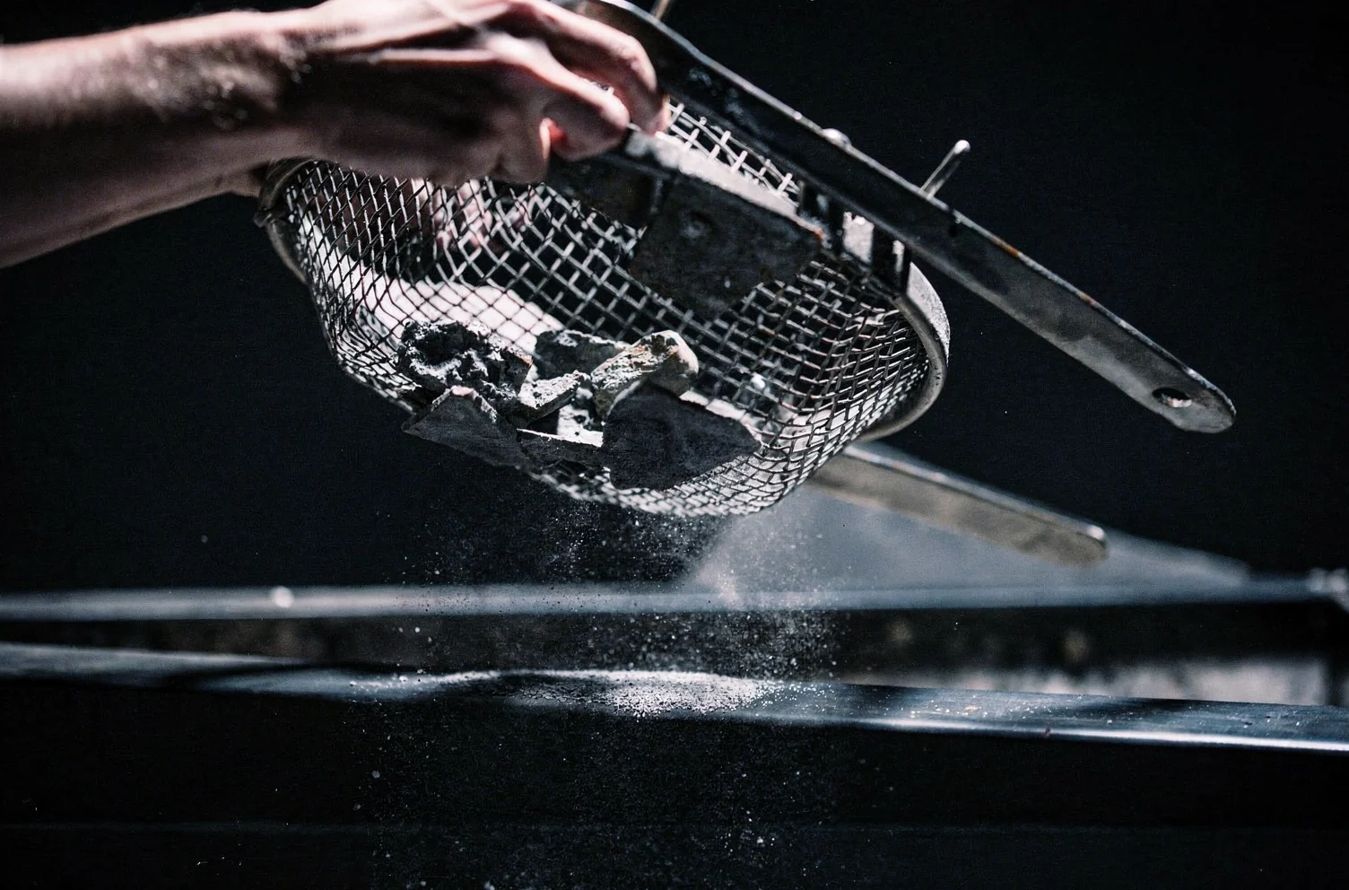 Two hands tilting a worn steel mesh sieve over a bench, coarse pieces caught in the mesh while fine dust falls through